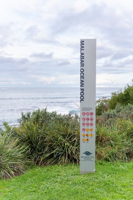 A tall, vertical metal sign displaying the words 'MALABAR OCEAN POOL' in black letters on a white background, positioned on a grassy area near the coast. The sign features various warning symbols in r