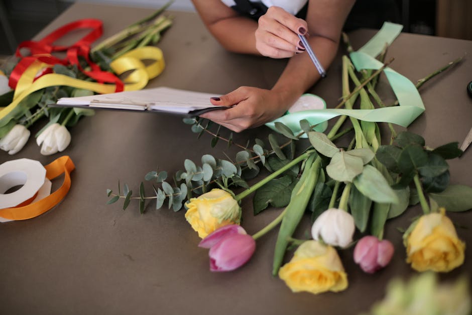 A florist's workspace featuring a woman holding a notepad and pen, surrounded by partially assembled floral arrangements. The table is scattered with fresh flowers including yellow and pink tulips, wh
