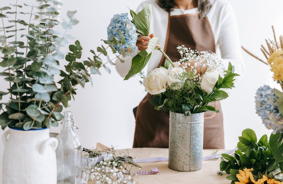 A professional florist at Clapham Florist, dressed in a brown apron over a white shirt, arranging a floral bouquet with white roses, baby's breath, and lush green foliage in a cylindrical metallic vas