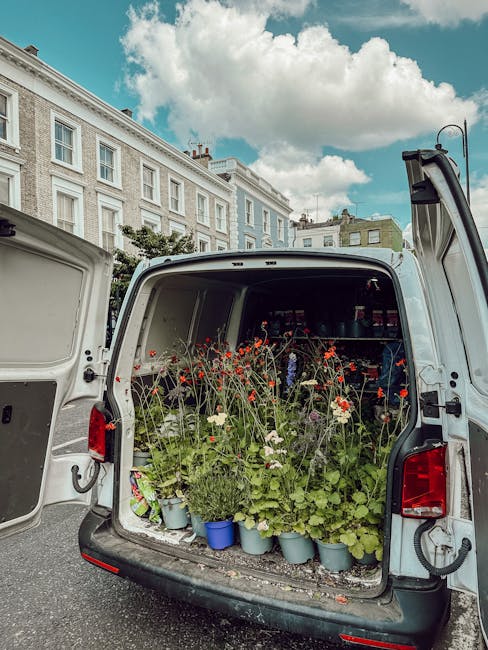 The open rear of a white delivery van filled with various potted flowers and plants, including red poppies, white daisies, and green foliage, arranged on shelves and in lightweight plastic pots. The f
