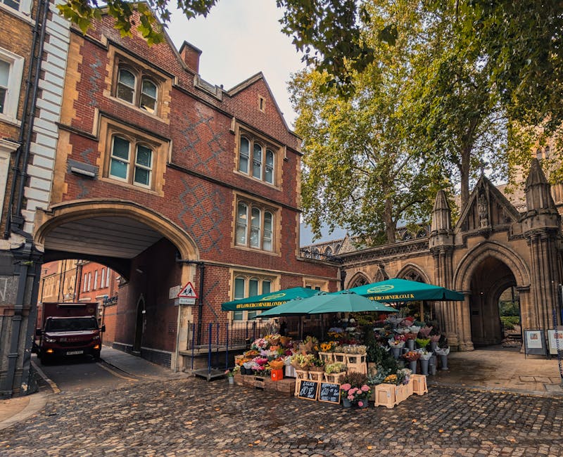 A vibrant outdoor flower stall situated beneath a green umbrella near an arched historic building entrance in Clapham. The stall is filled with a variety of fresh flowers, including pink roses, white